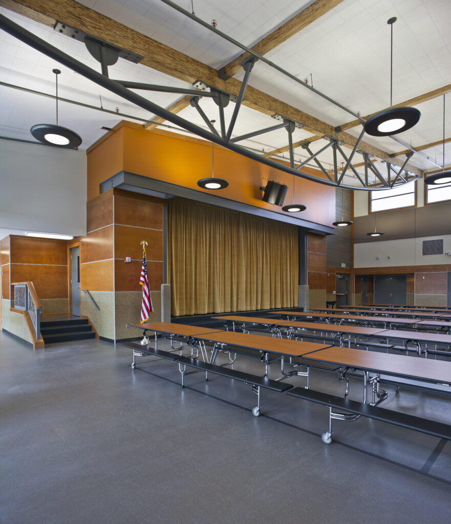Large school cafeteria with foldable tables and a stage area before renovation and modernization upgrades.